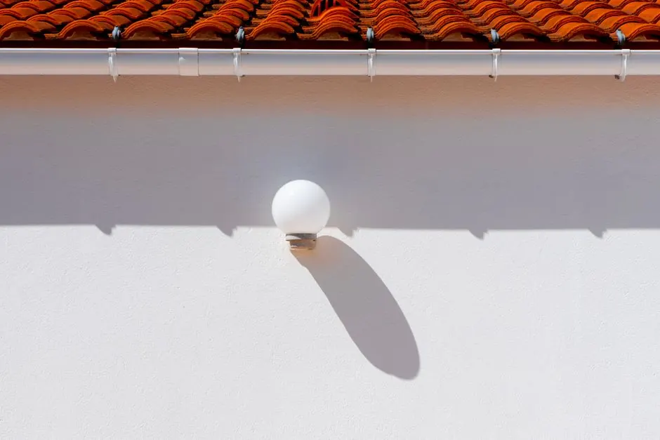 A minimalist outdoor setting showcasing a wall lamp casting a shadow beneath a red tiled roof.